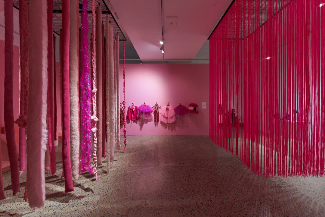 Installation view of a contemporary art exhibition dominated by shades of pink. Long, textured fabric and yarn pieces hang from the ceiling on the left, while a dense curtain of pink cords creates a partition on the right. Pink garments and sculptural textile works are displayed on the back wall in a softly lit gallery with a terrazzo floor.